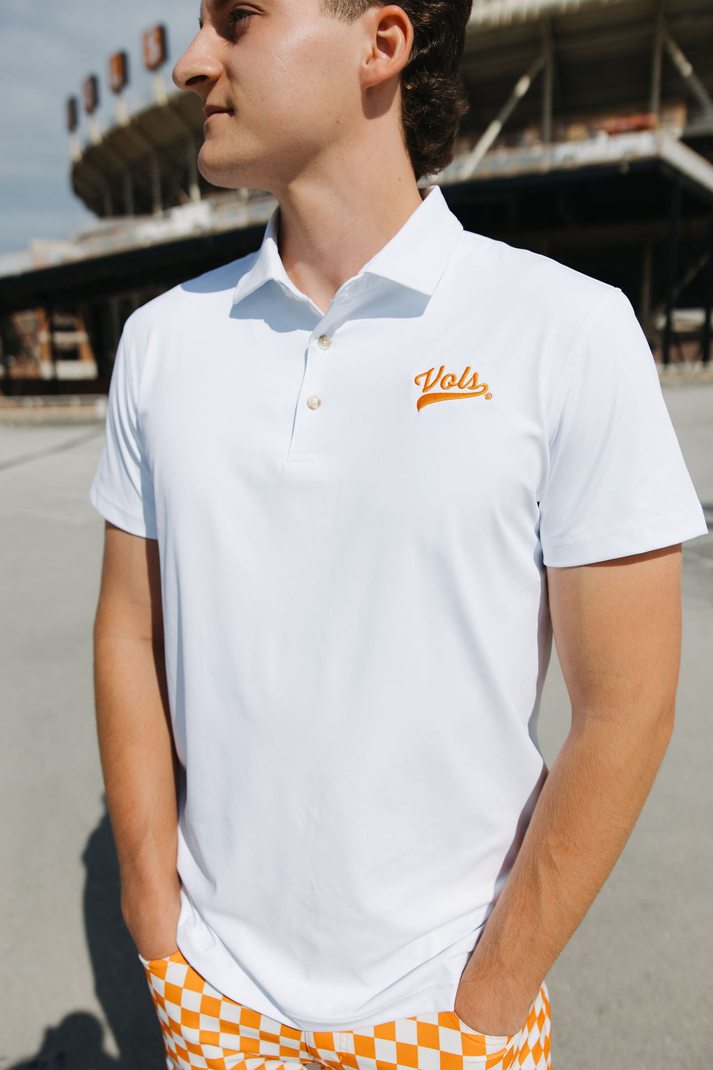 A person wearing a white polo shirt with Vols embroidered in orange on the chest stands outdoors near a sports stadium, with hands in orange and white checkered pants pockets.