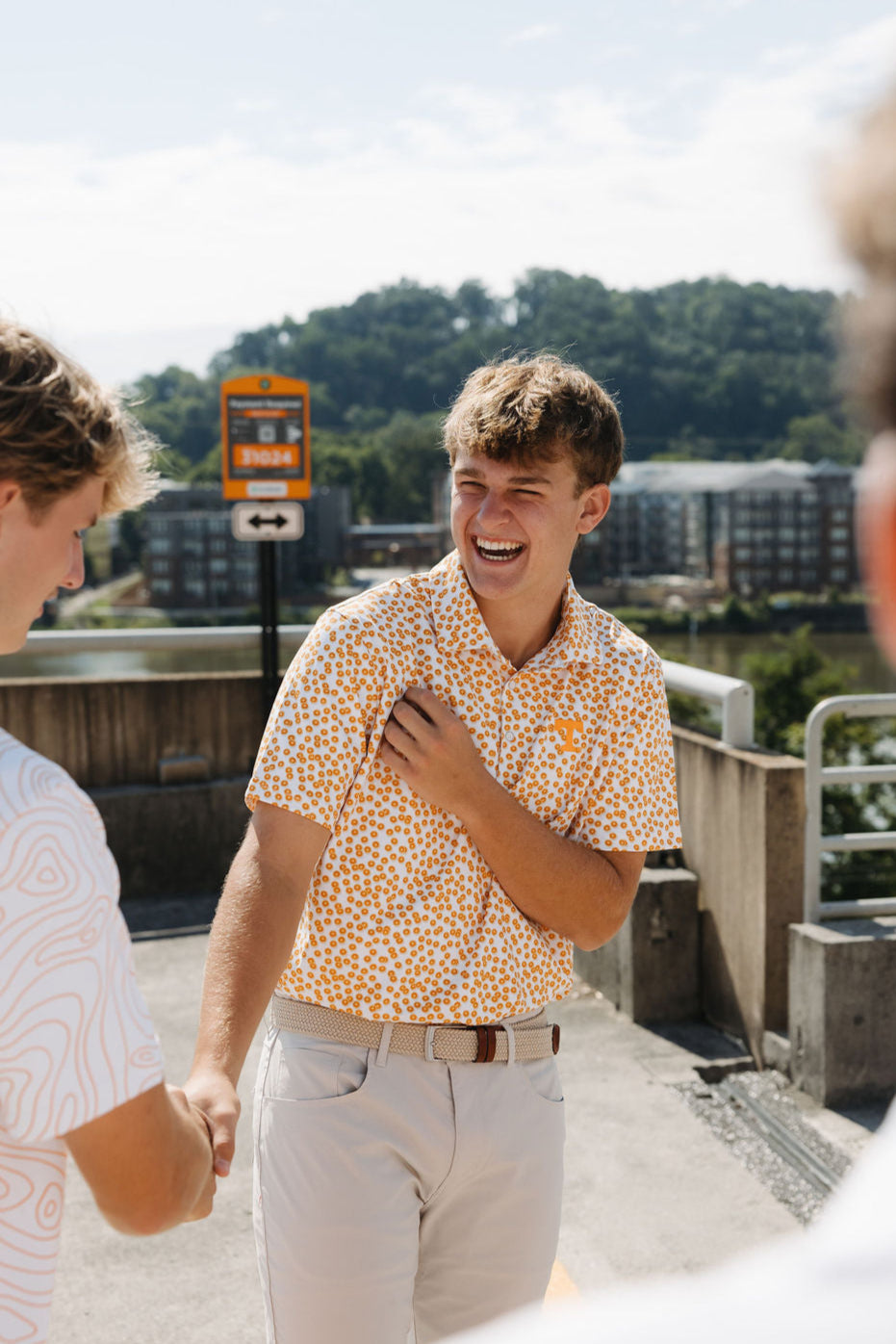 A young man wearing a patterned short-sleeve shirt and light pants laughs while shaking hands with another person outdoors in a sunny parking lot, with trees and buildings in the background.