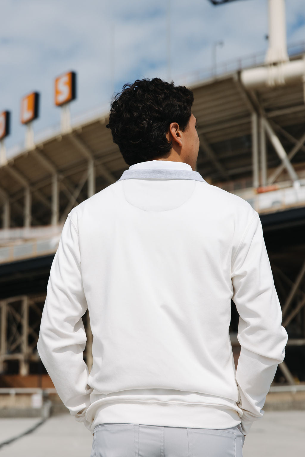 A man with curly brown hair, dressed in a white long-sleeve shirt and light pants, stands outdoors with his back to the camera in front of a stadium with large signs in the background.