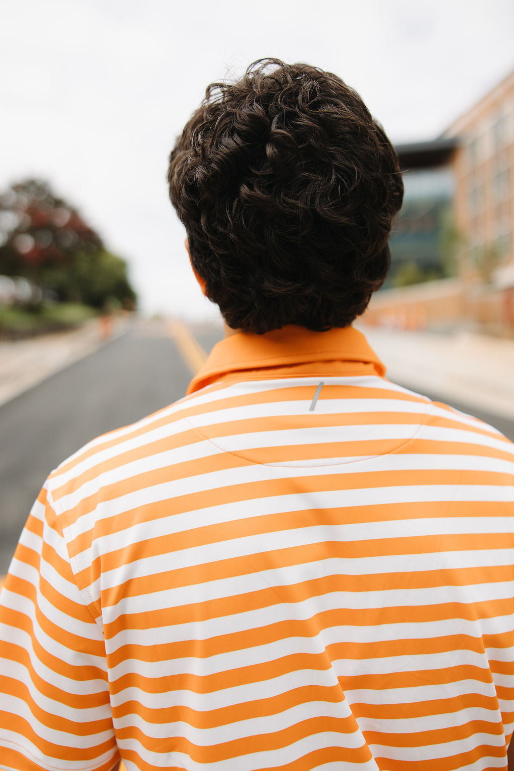 A person with short, curly dark hair stands outdoors, facing away from the camera. They are wearing an orange and white striped collared shirt. The background shows a road and blurred buildings and trees.