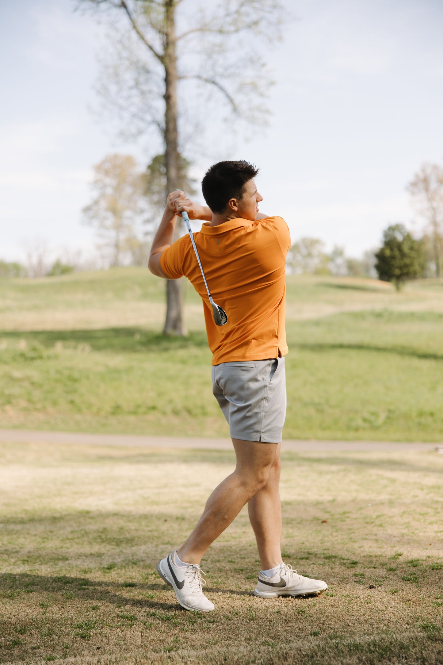 A man in an orange polo shirt and gray shorts swings a golf club on a grassy course, with trees and a blue sky in the background.