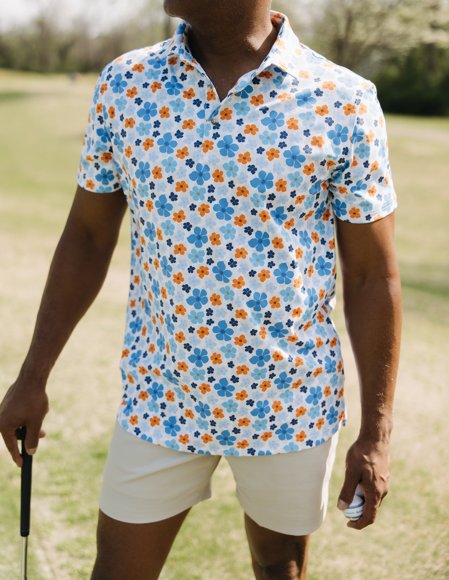 A person wearing a white polo shirt with blue and orange floral patterns and beige shorts stands on a golf course, holding a golf club and a golf ball. The scene is outdoors on a sunny day.