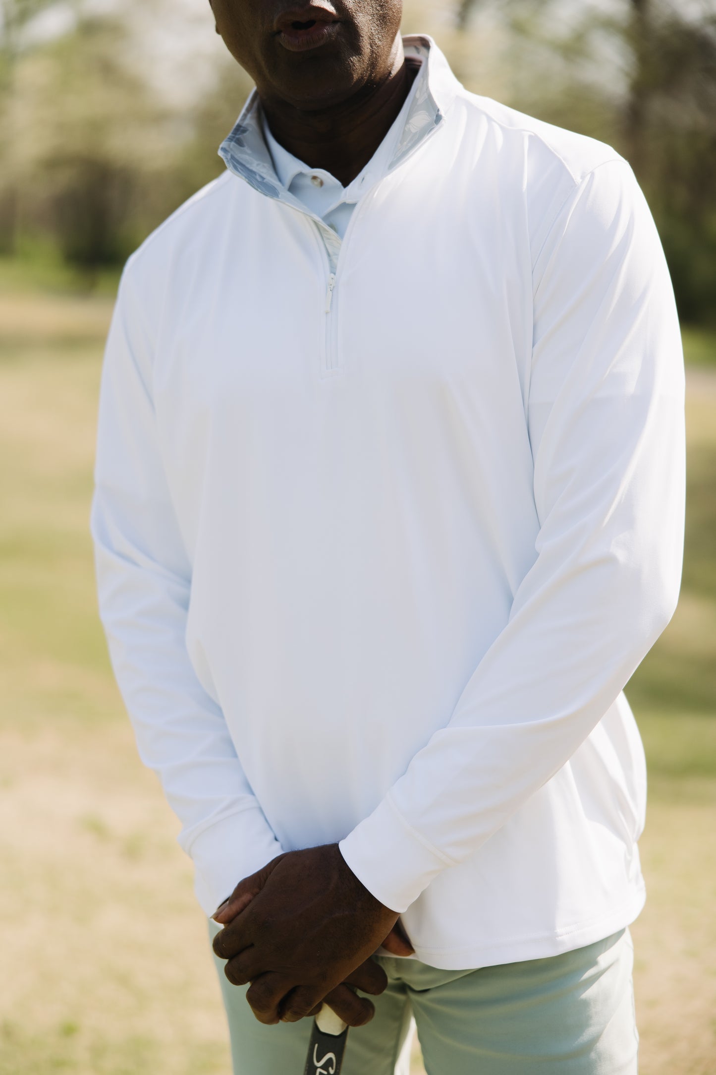 A man wearing a white quarter-zip pullover and light pants stands outdoors on grass, holding a golf club with both hands; his face is partially out of frame.