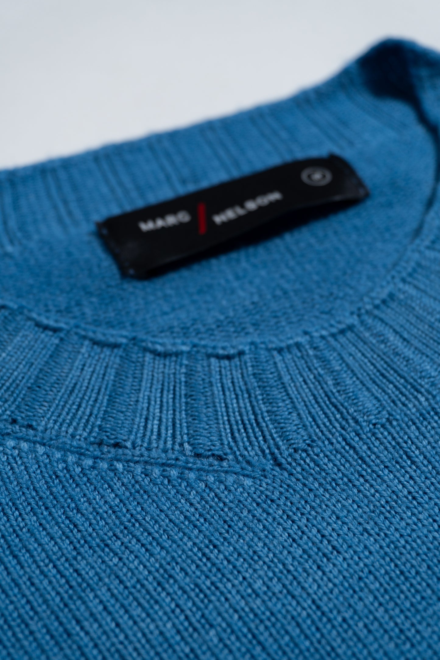 Close-up of a blue knitted sweater with a ribbed neckline, showing a black label with white text and a red slash, partially reading MADE / MELDUN. The texture of the fabric is clearly visible.