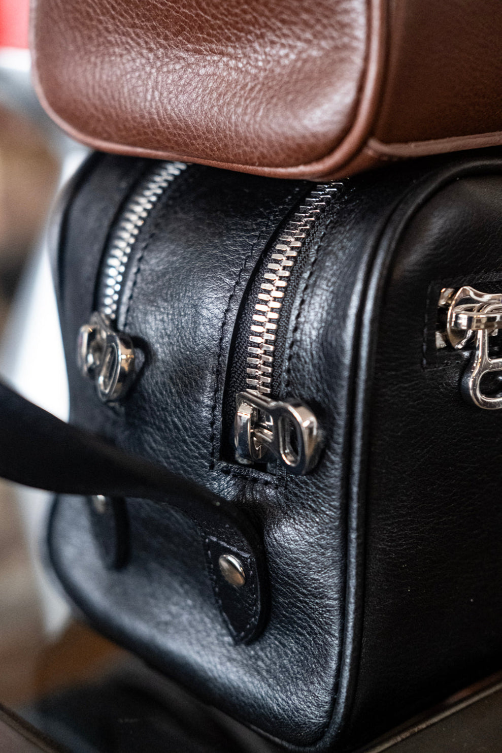 Close-up of a black leather bag with silver zippers stacked underneath a brown leather item, highlighting the textures and details of the materials and hardware.