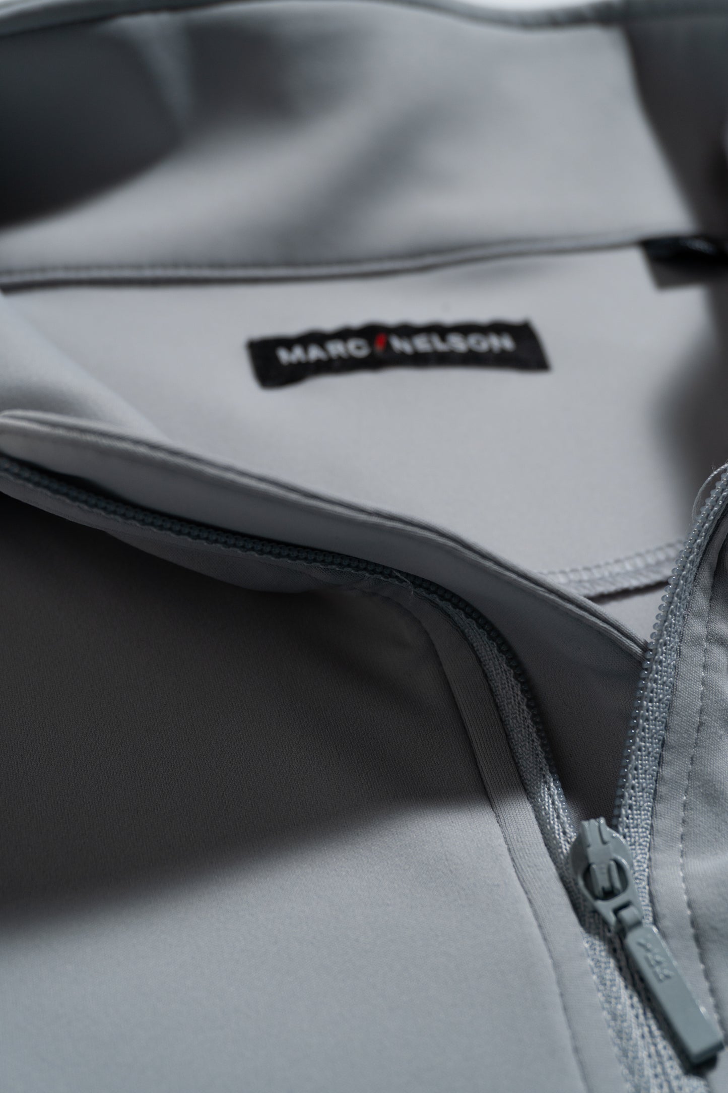 Close-up of a light gray zip-up garment, partially unzipped to reveal a black label with the text MARC NELSON stitched inside the collar. The fabric appears smooth and slightly reflective.