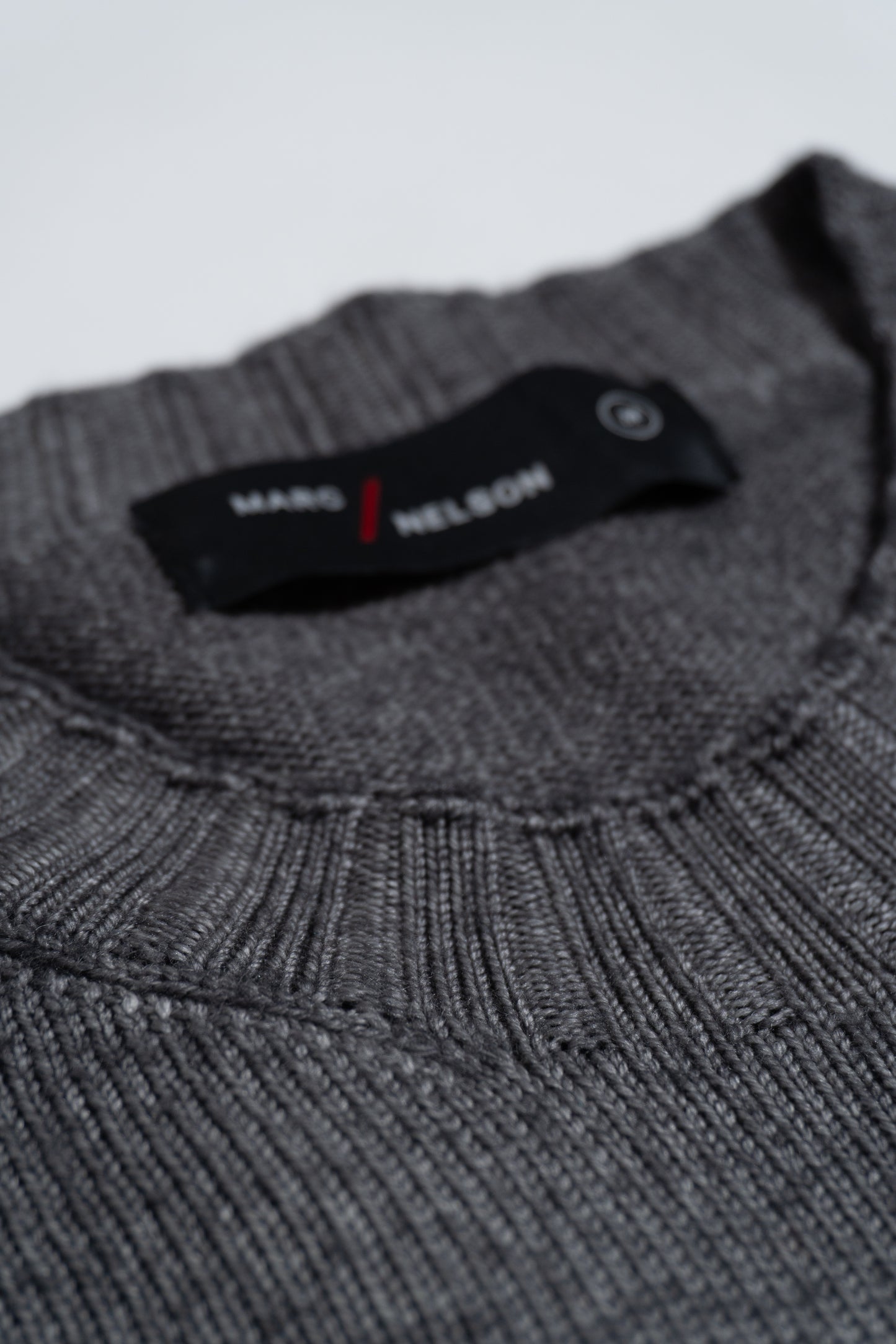 A close-up of a folded gray knitted sweater with a visible black clothing label that reads MARS / NELSON in white and red text. The sweater’s ribbed texture and neckline are prominent.