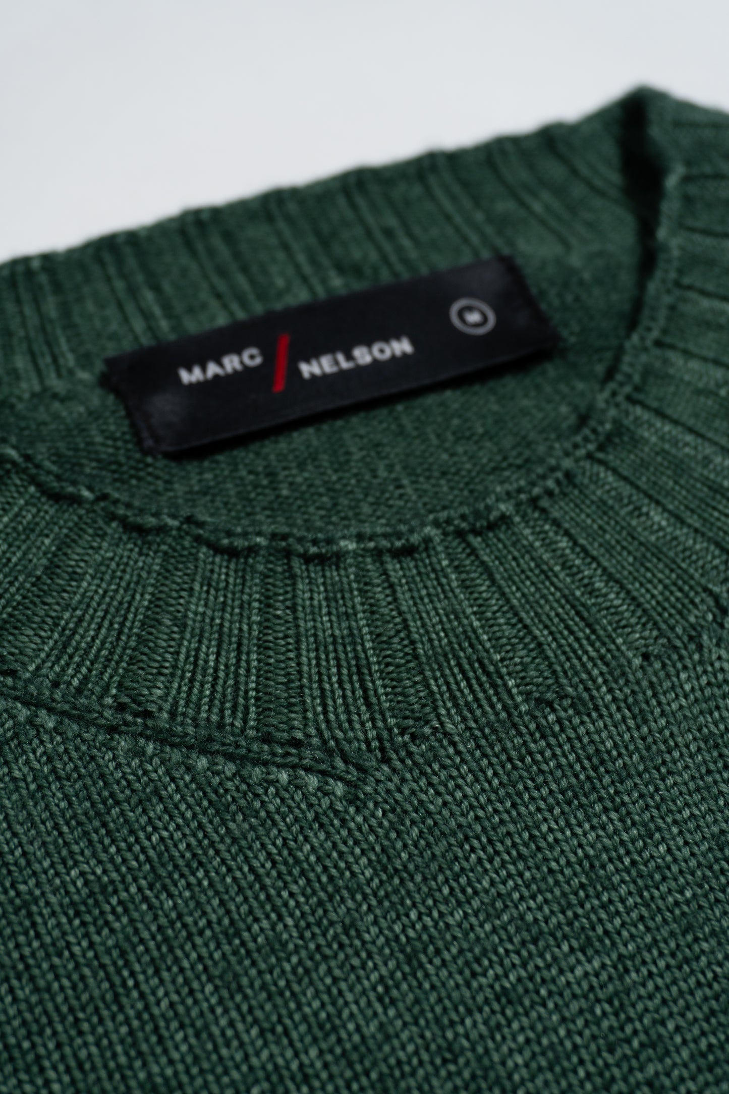 Close-up of a green knitted sweater with a ribbed neckline. A black label on the inside collar reads MARC / NELSON in white and red text.