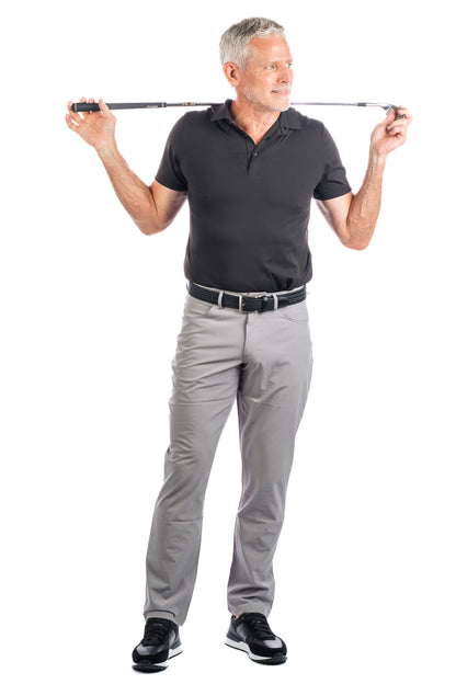 Photo of model wearing a black golf polo and concrete five pocket pants on a white background and holding a golf club on his shoulders. 