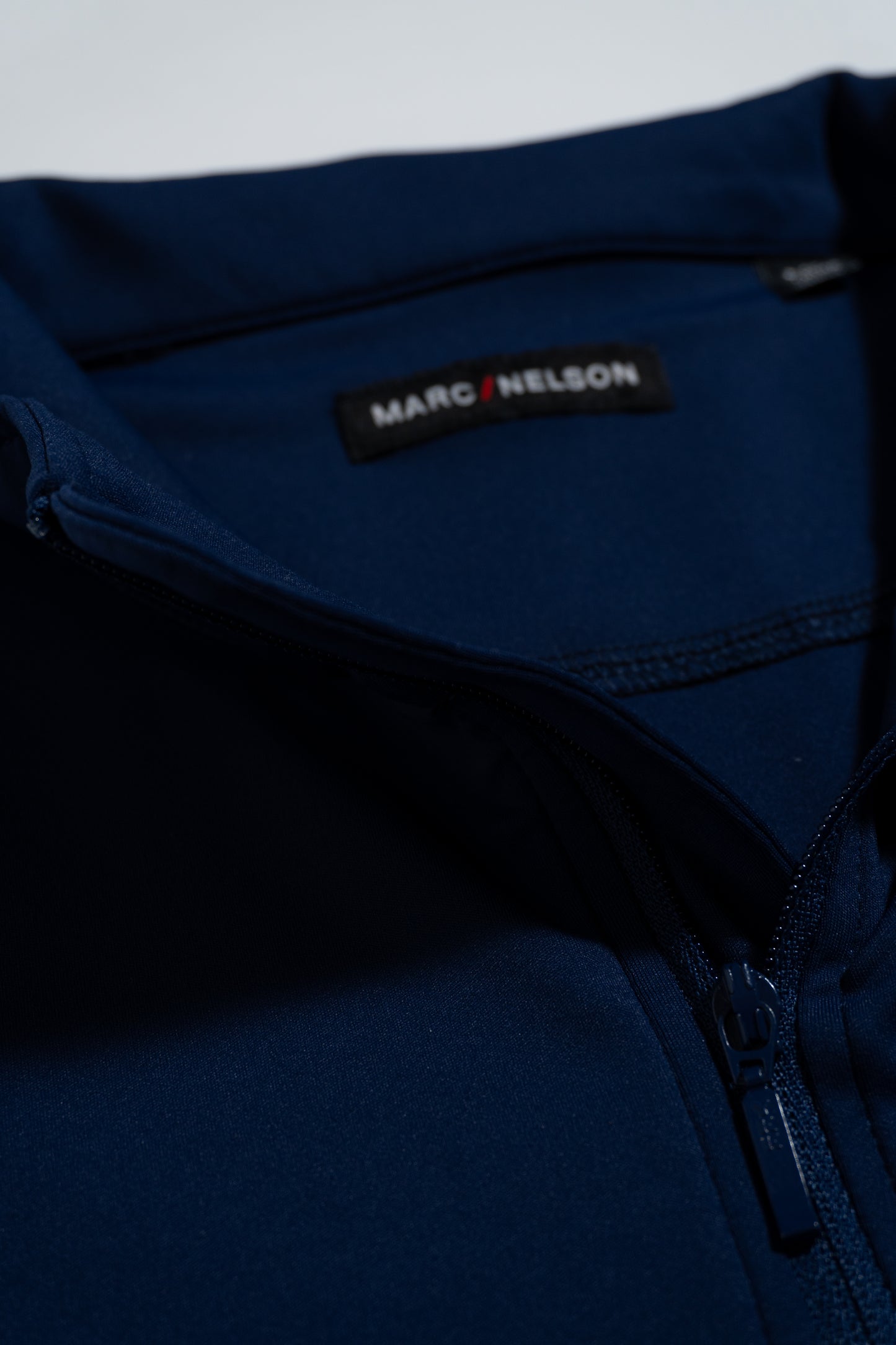 Close-up of a navy blue zip-up garment with a visible MARC NELSON label on the inside of the collar. The zipper and stitching details are clearly shown.