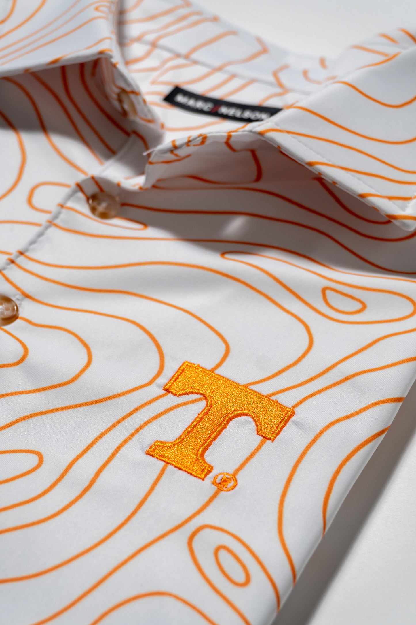 A white button-up shirt with orange contour line patterns and an embroidered orange T logo on the chest, shown folded neatly on a white surface.