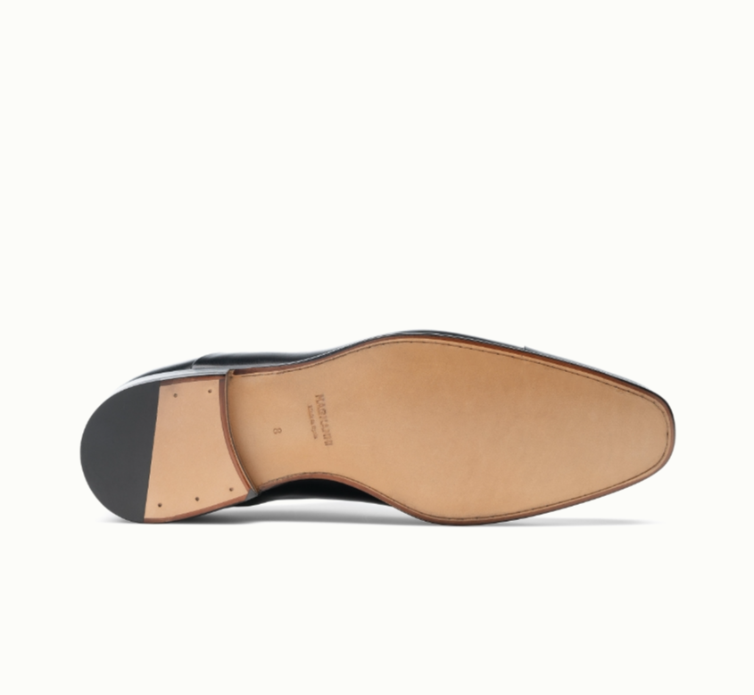 The image shows the sole of a black formal dress shoe lying on its side. The sole is tan leather with visible stitching and a black heel, displaying craftsmanship details.
