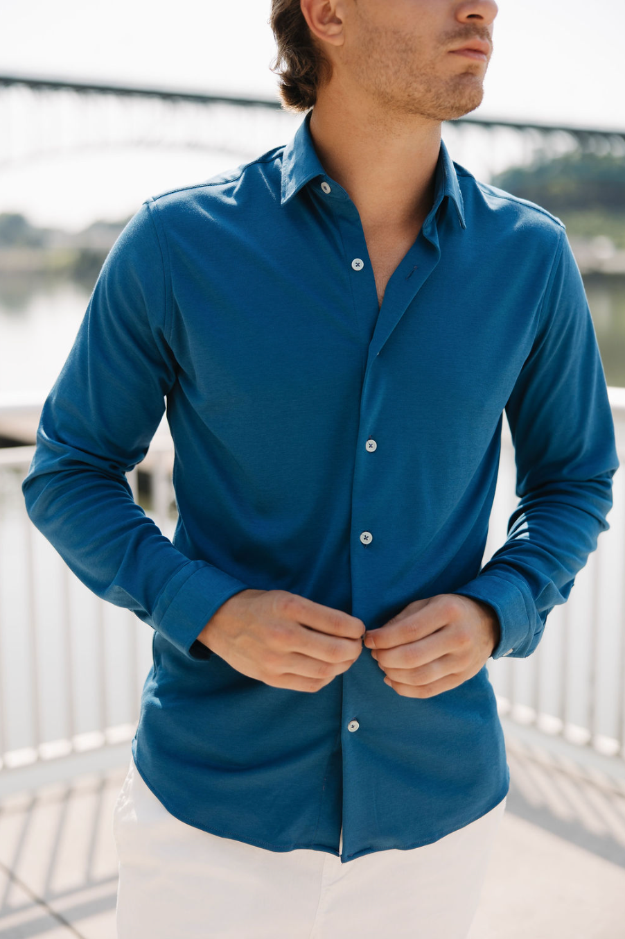 A man in a blue button-up shirt and white pants buttons his shirt while standing outdoors near a railing, with a blurred bridge and water in the background. His face is partially out of frame.