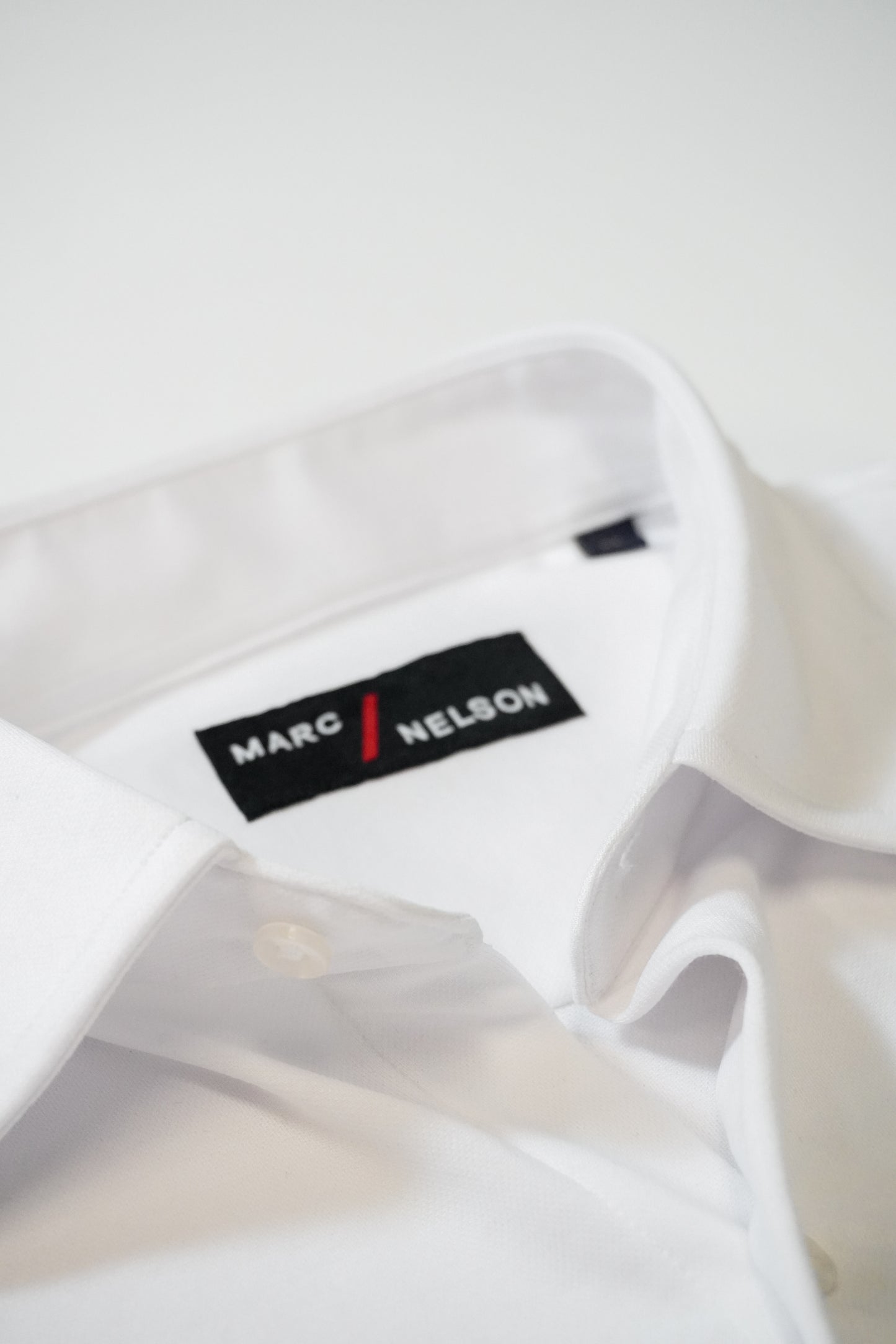 A close-up of a neatly folded white dress shirt with the collar visible and a black label that reads MARC NELSON stitched inside the collar.