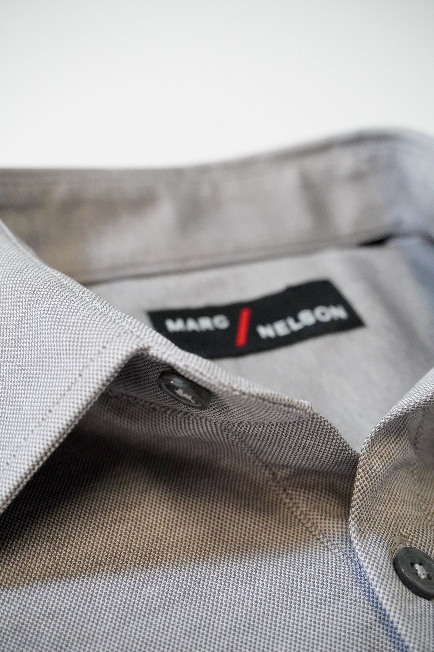 Close-up of a light gray dress shirt with black buttons, focusing on the collar and a black label that reads MARC NELSON in white and red text.