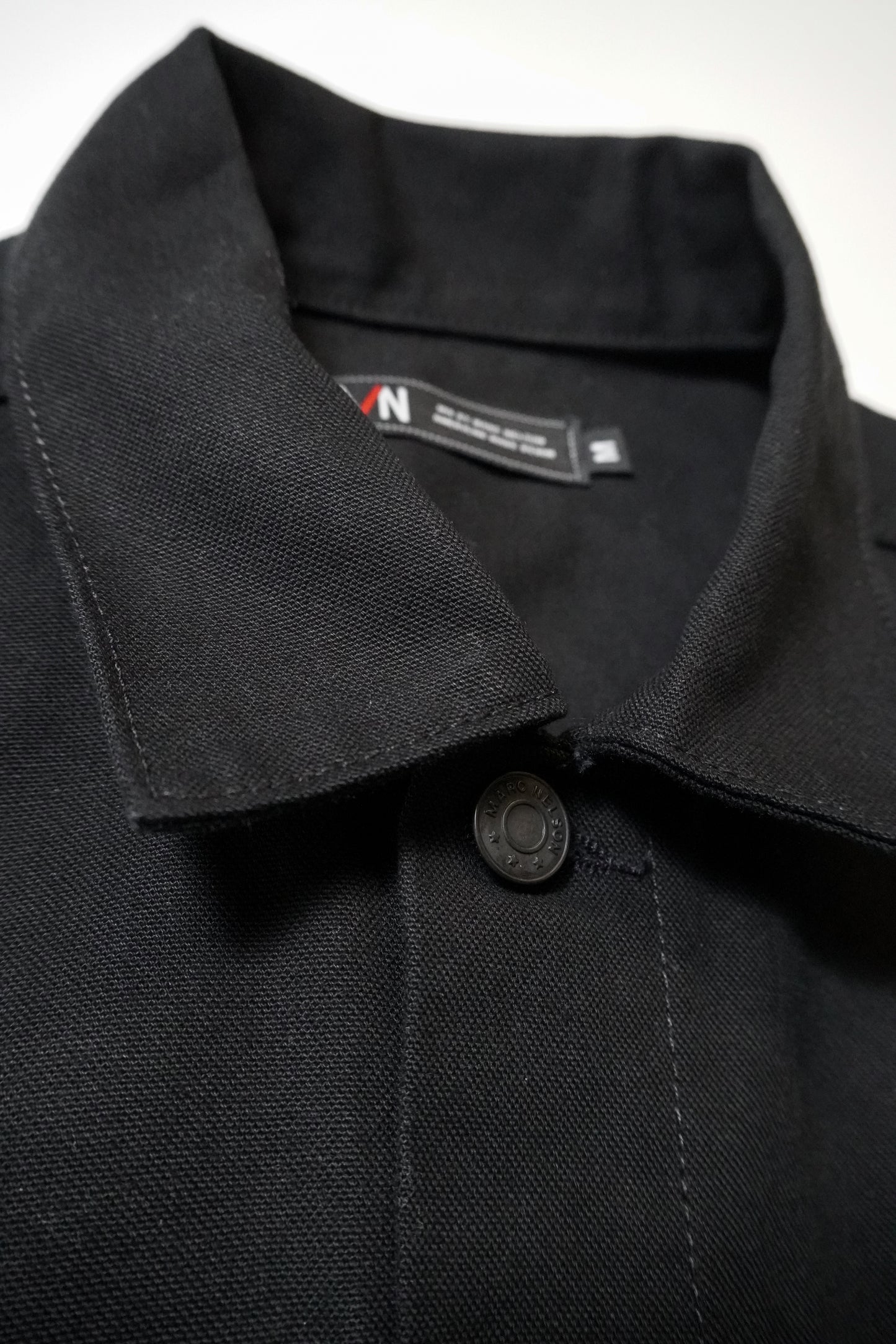 Close-up of a black collared jacket made from textured fabric, showing the top button fastened and a visible clothing label inside the collar.