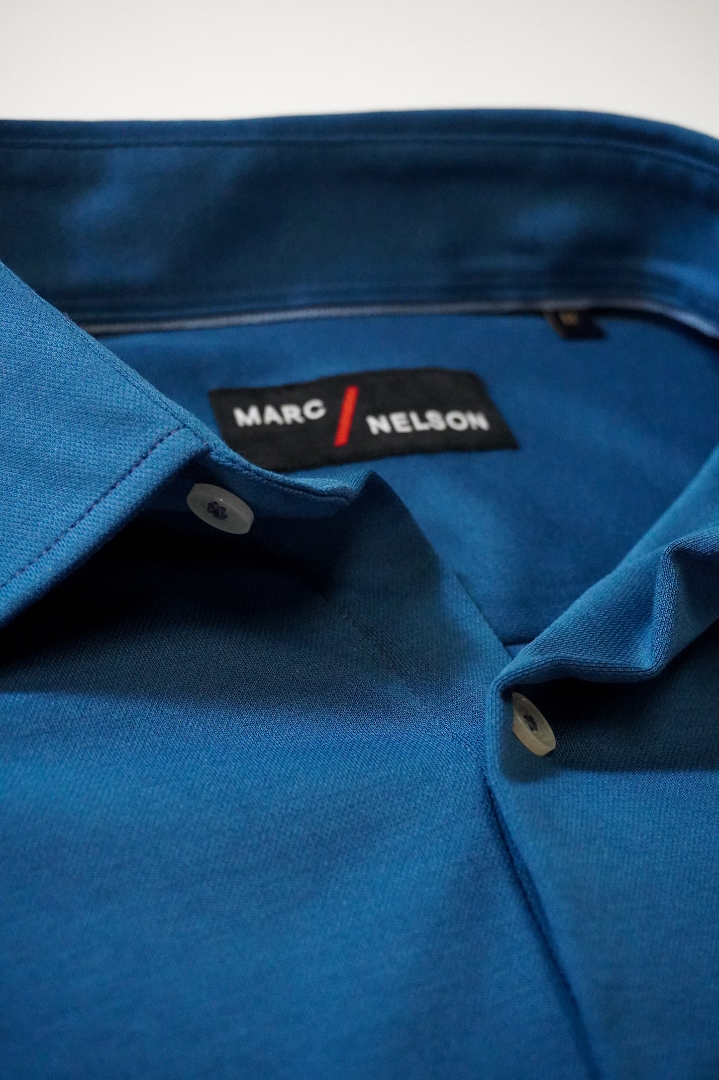 Close-up of a blue collared shirt with white buttons, showing the black label inside the collar that reads MARC / NELSON in white text.