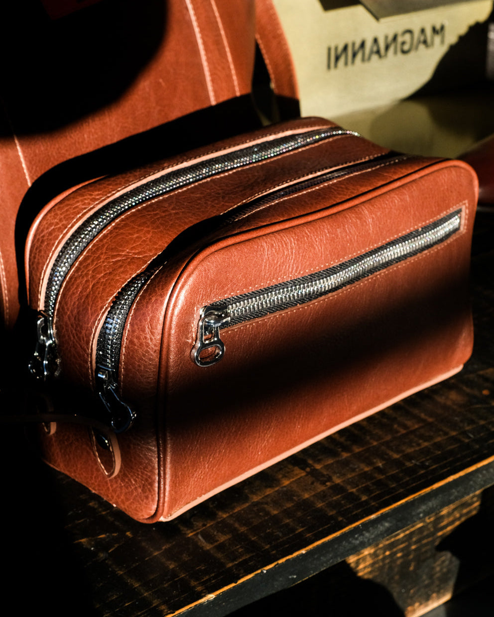 A brown leather toiletry bag with silver zippers sits on a wooden surface, partially in shadow. In the background, part of a tan bag with the word MAGNANI is visible.