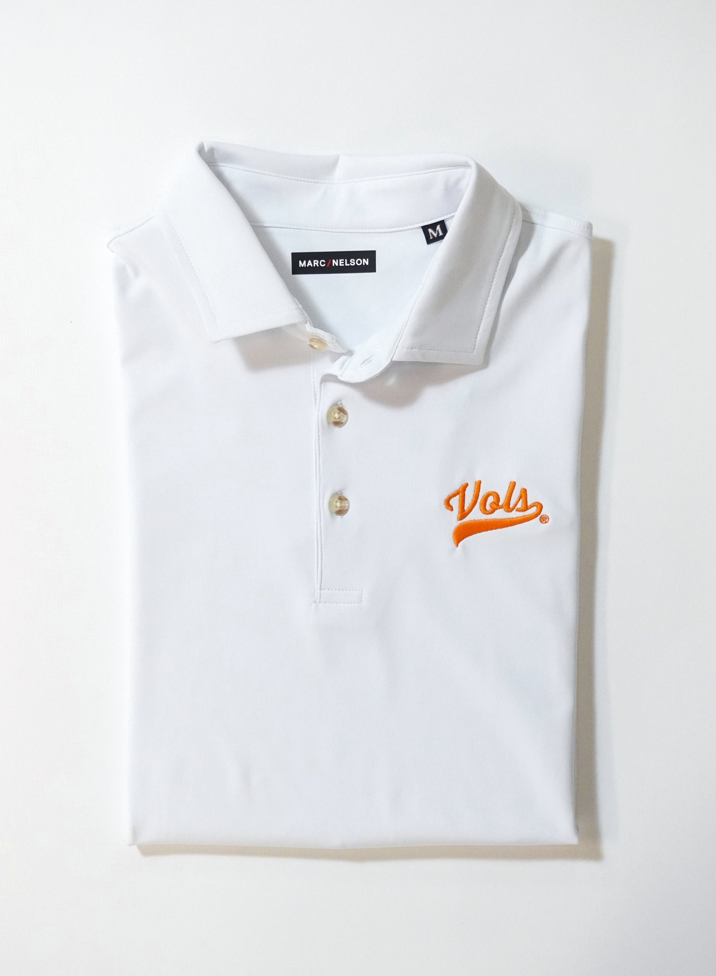 A folded white polo shirt with three buttons and an orange Vols logo on the chest, featuring a Marc Nelson label and a size M tag inside the collar.