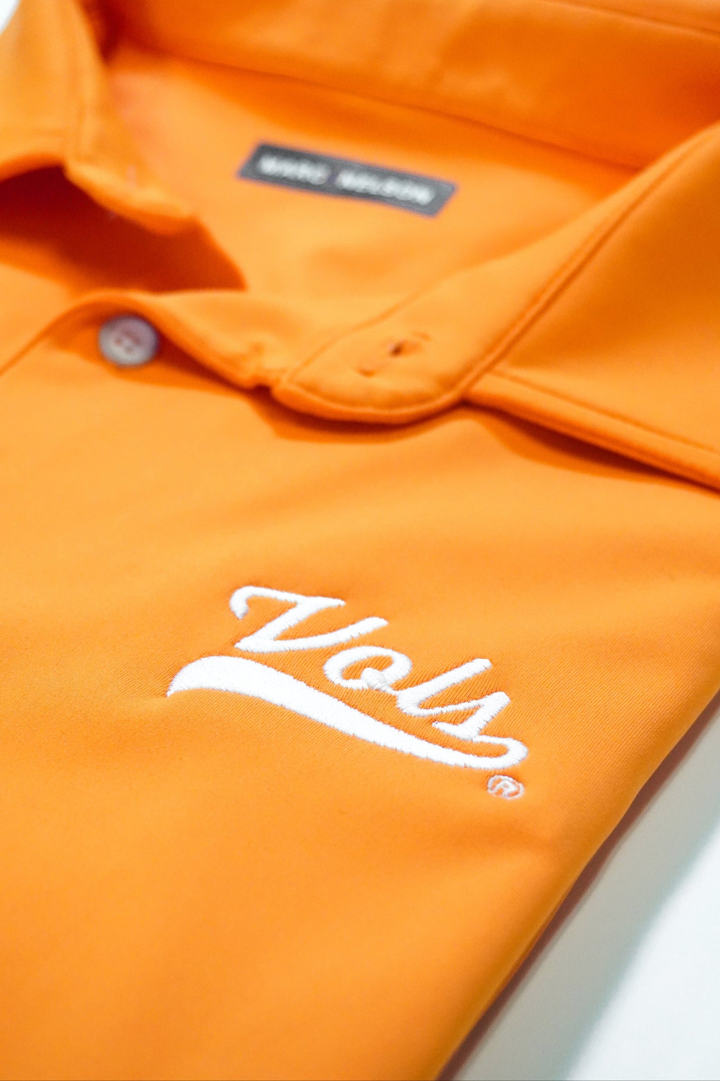 Close-up of an orange collared shirt with a white Vols logo embroidered on the chest. A button and part of the collar are visible, as well as a blurred label inside the shirt.