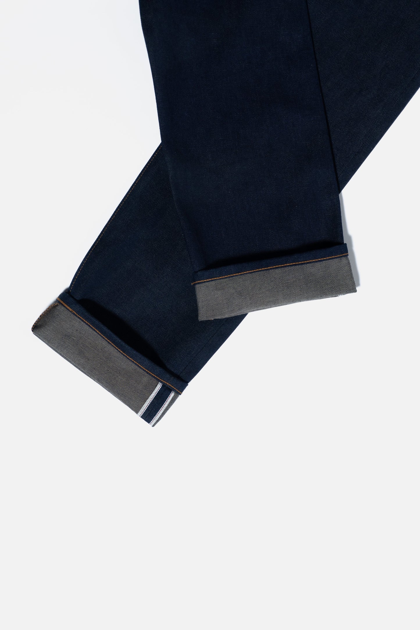 Two pairs of dark blue denim jeans with the cuffs neatly rolled up, displaying the contrasting inner fabric and striped selvedge edges, set against a plain white background.
