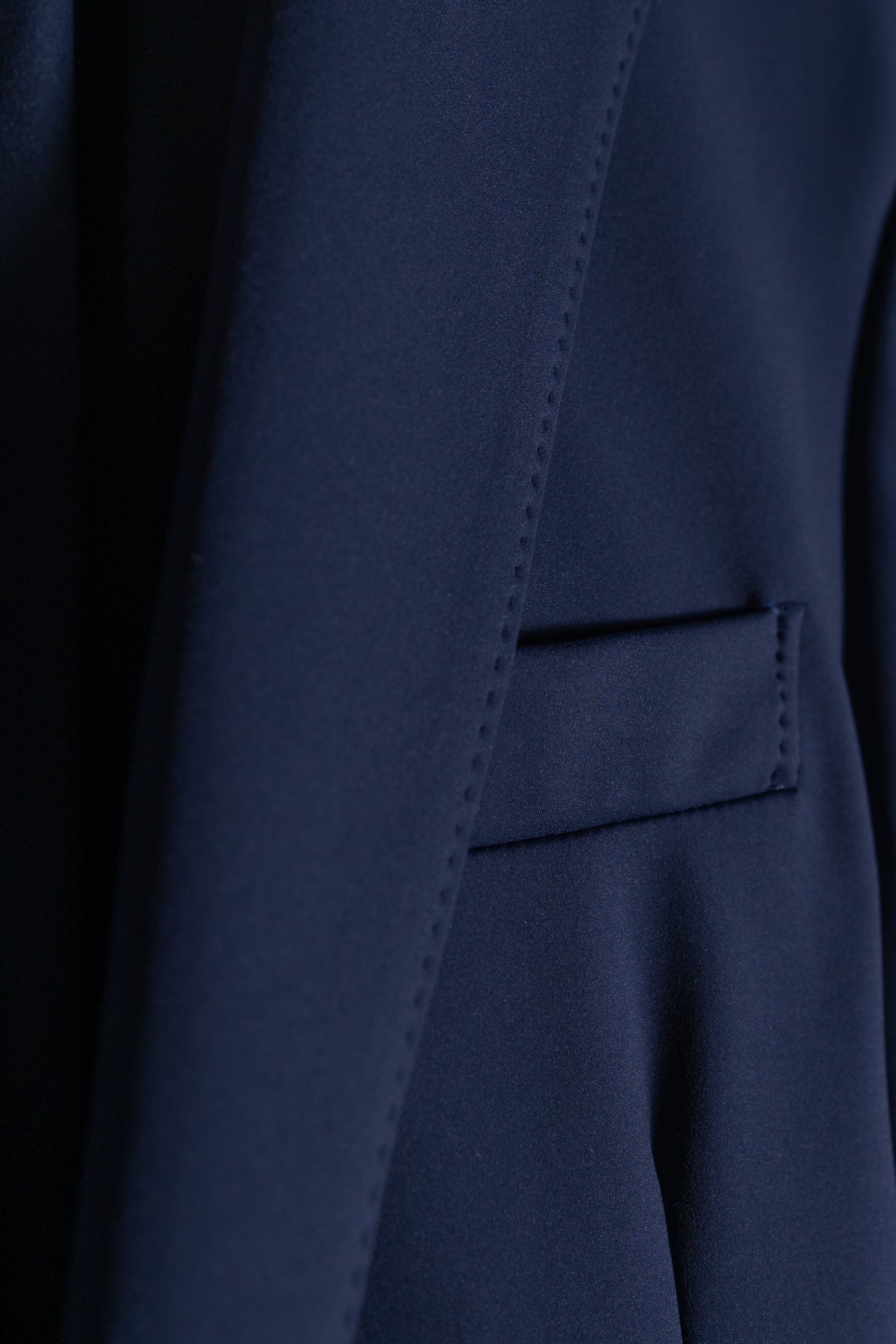 Close-up of a navy blue blazer, showing a lapel and a pocket with a flap, highlighting the smooth texture and stitching details of the fabric.