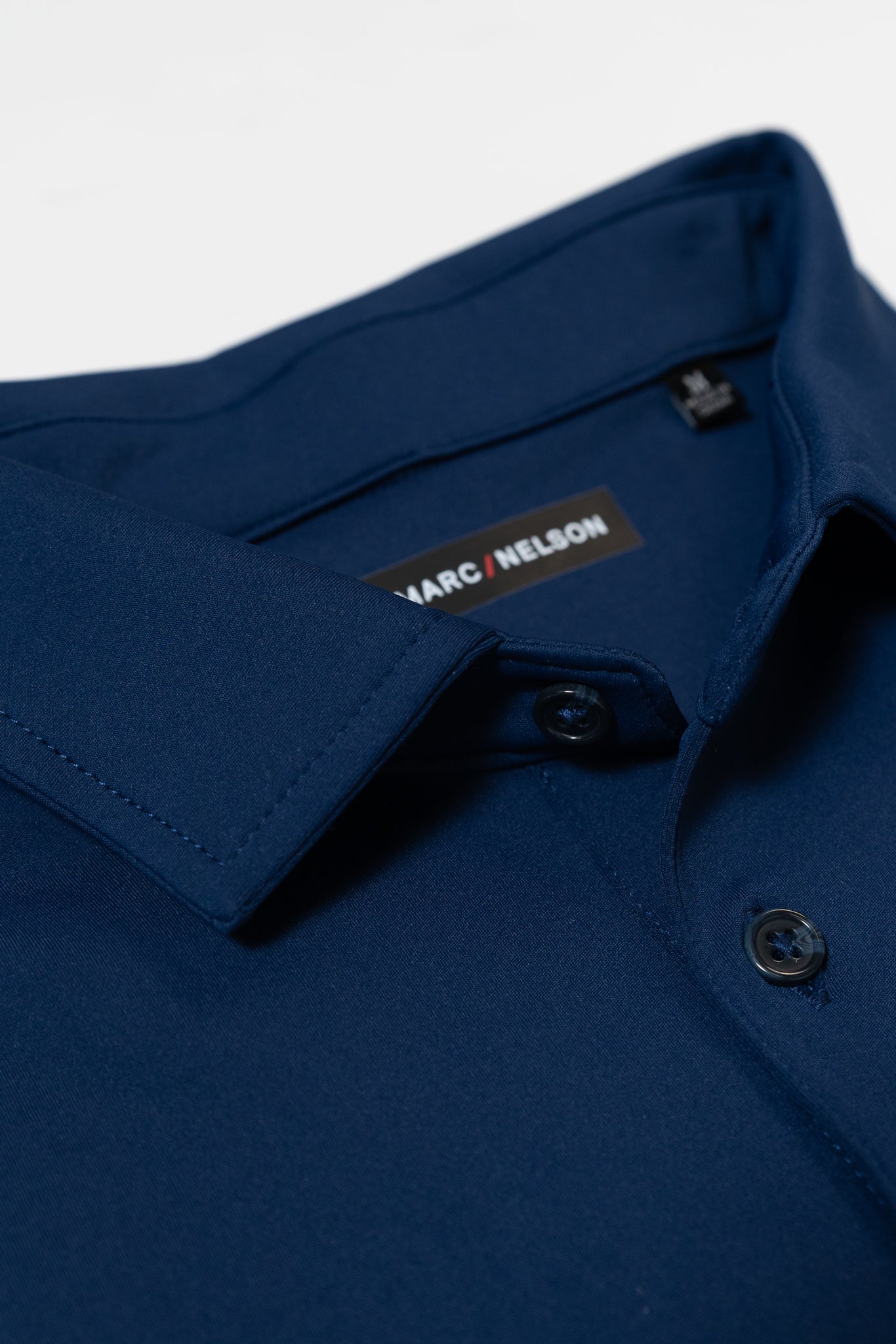 Close-up of a navy blue button-up shirt with a pointed collar and two visible buttons. The inside label reads MARC / NELSON on a black background.