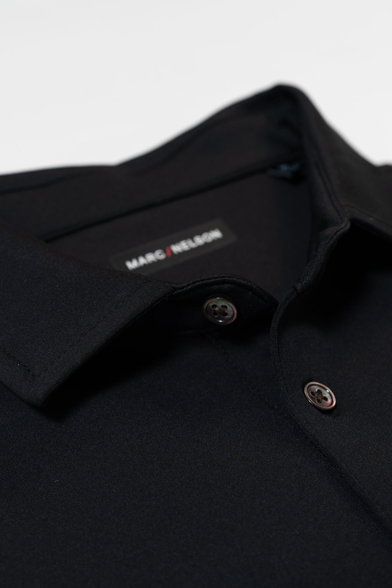 Close-up of a black collared shirt with two buttons visible. The shirts label, which reads MARC NELSON, is partially visible inside the collar. The fabric appears smooth and neatly pressed.