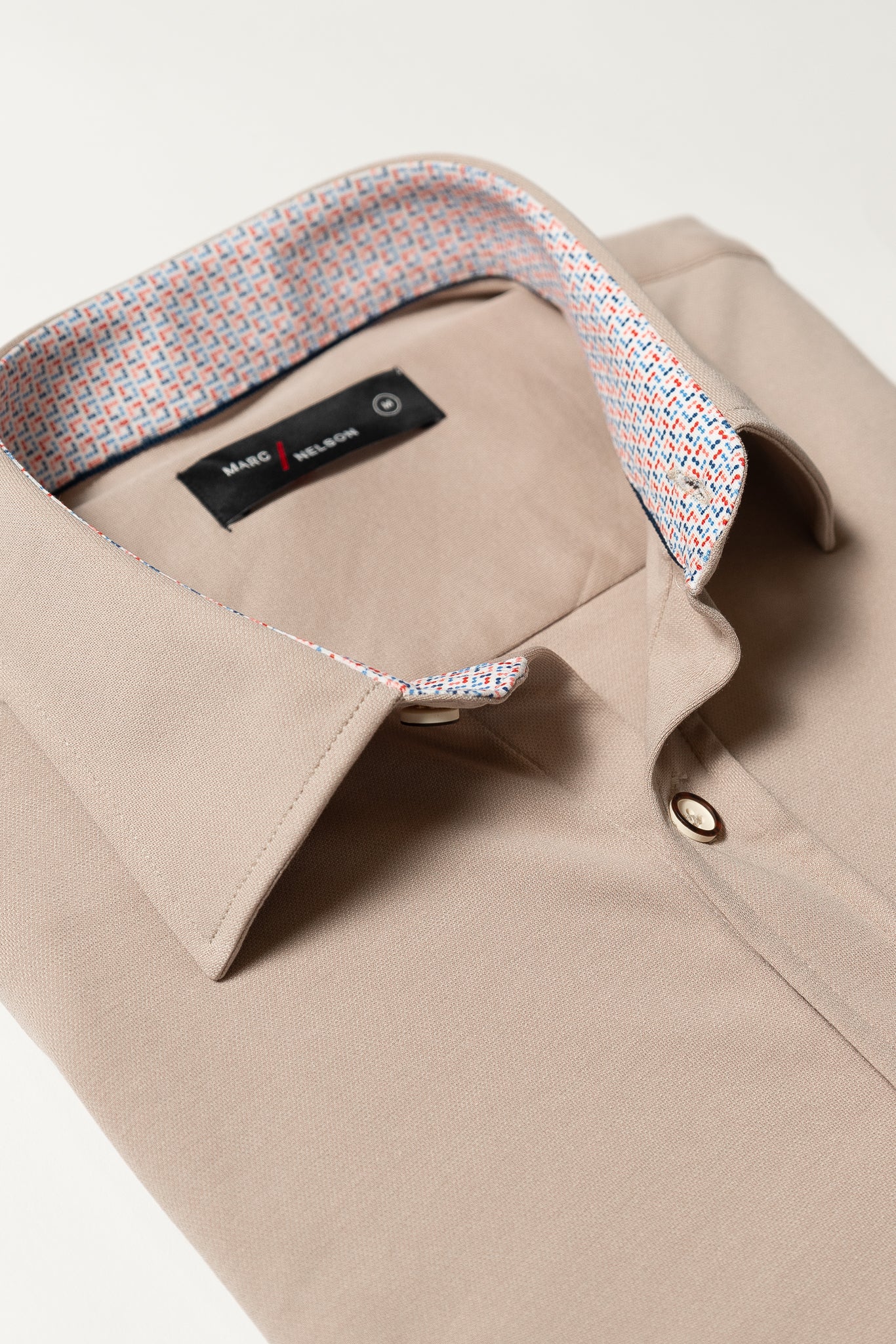 A neatly folded beige dress shirt with a patterned inner collar featuring red, blue, and white designs. The shirt has a button front and a black label inside the collar.