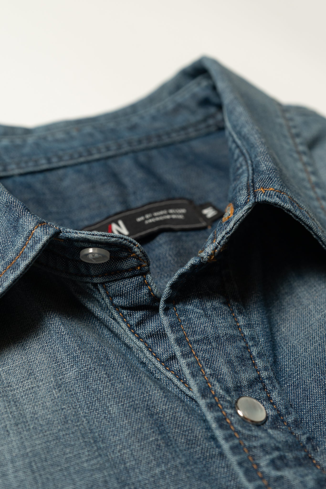 Close-up of a blue denim shirt with metal snap buttons and visible stitching, focusing on the collar and top part of the shirt against a light background.
