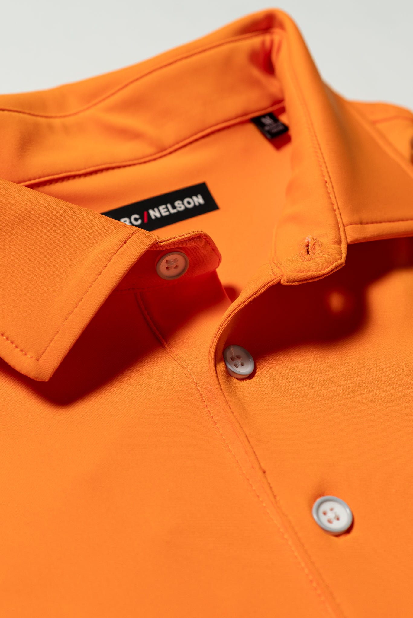Close-up of an orange collared shirt with white buttons. The shirt label inside the collar reads BC/NELSON. The fabric appears smooth and neatly stitched.