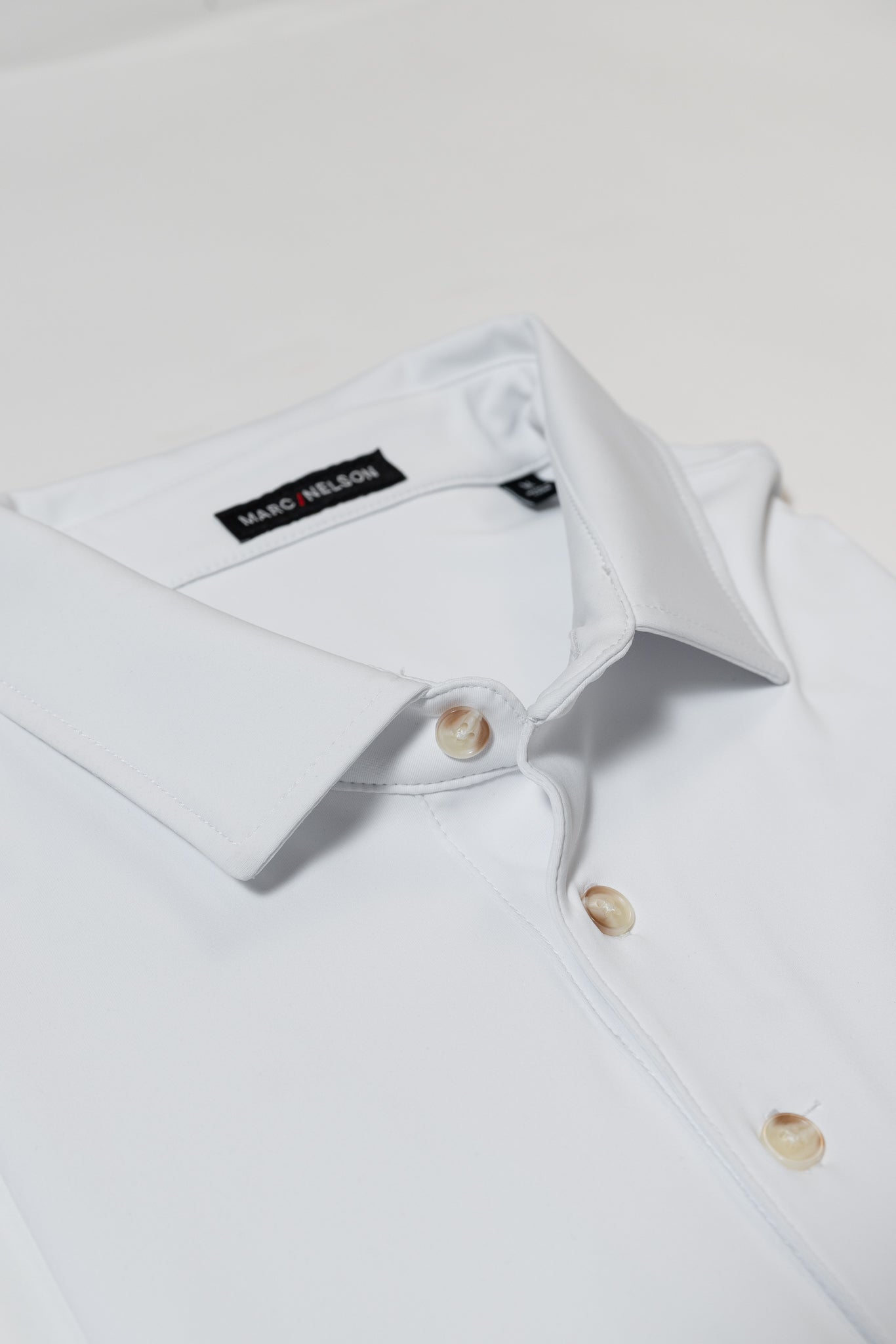A close-up of a neatly folded white dress shirt with ivory buttons and a visible black Marc Nelson label on the collar, set against a plain white background.