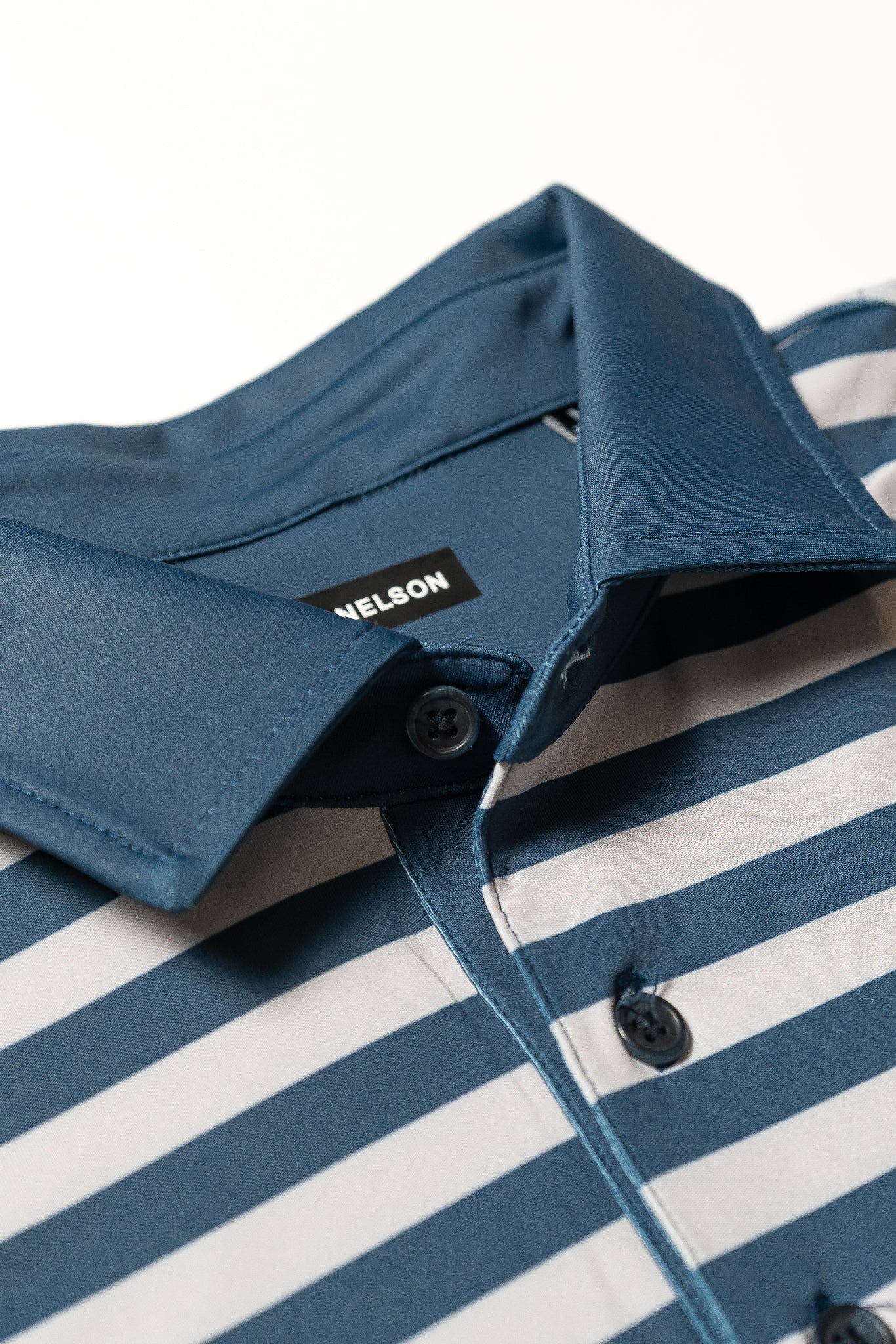Close-up of a blue and white horizontal striped collared shirt with dark buttons. The shirt tag inside the collar reads NELSON. The fabric appears smooth and slightly shiny.