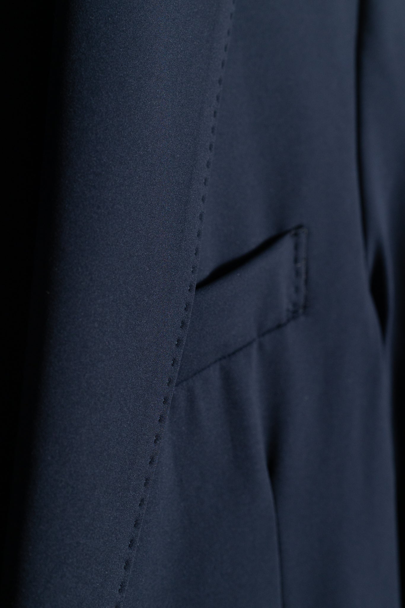 Close-up of a navy blue suit jacket, showing a neatly stitched pocket and part of the sleeve. The fabric appears smooth and formal.