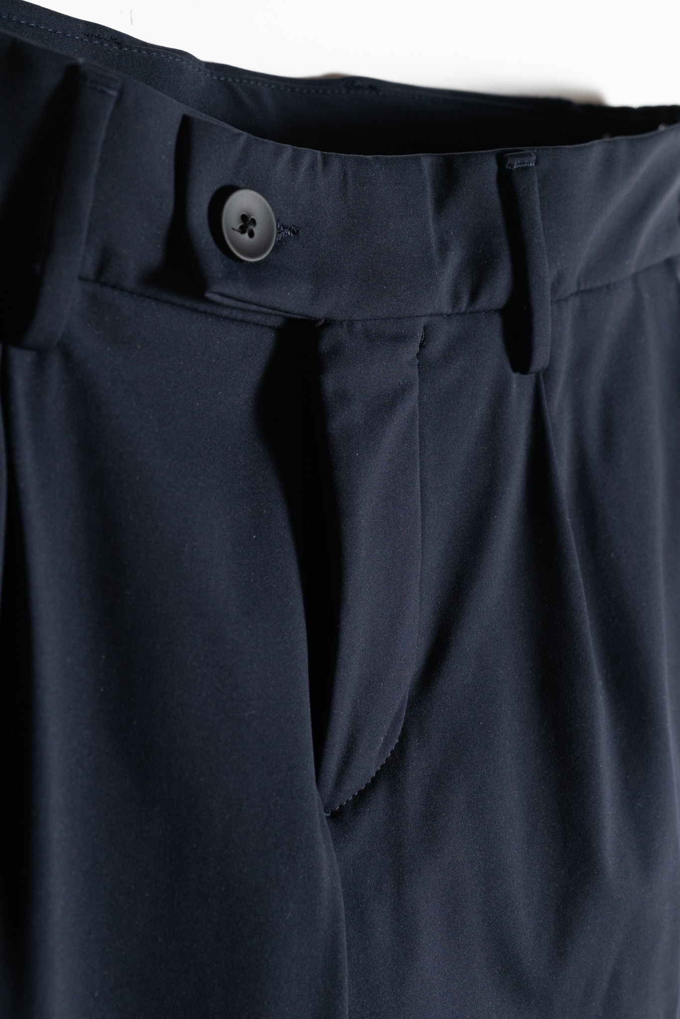 Close-up of the waistband and upper section of dark navy dress pants, showing a black button, belt loops, and a neatly tailored zipper fly.