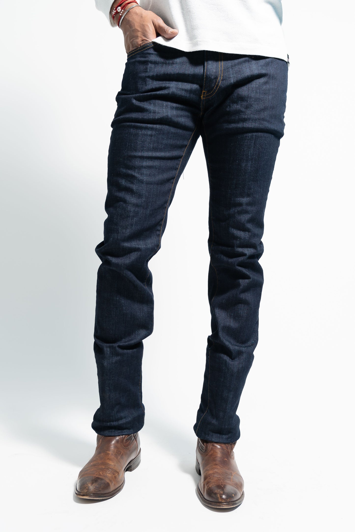 Man in slim straight dark wash jeans and brown boots standing on a white background.