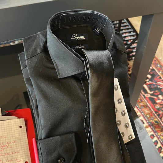 A neatly folded black dress shirt with a matching black tie placed on top, both labeled Lugano. Nearby are a white tie with a pattern and a notepad with graph paper and handwritten notes.