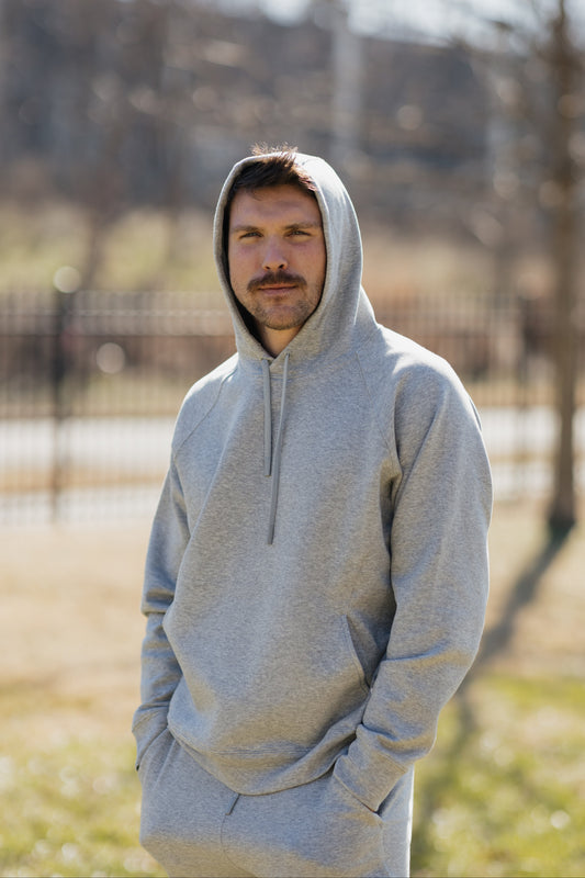 A man with short hair and a mustache stands outdoors wearing a light gray hoodie with the hood up, hands in his pockets, on a sunny day with a blurred background of trees and a fence.