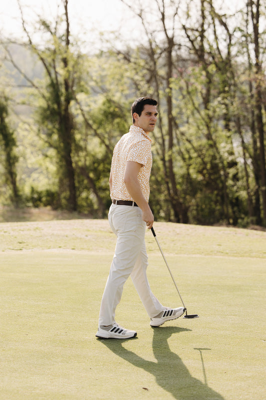 A man in a patterned short-sleeve shirt, light pants, and sneakers holds a golf club while walking on a green. Trees and sunlight are visible in the background.
