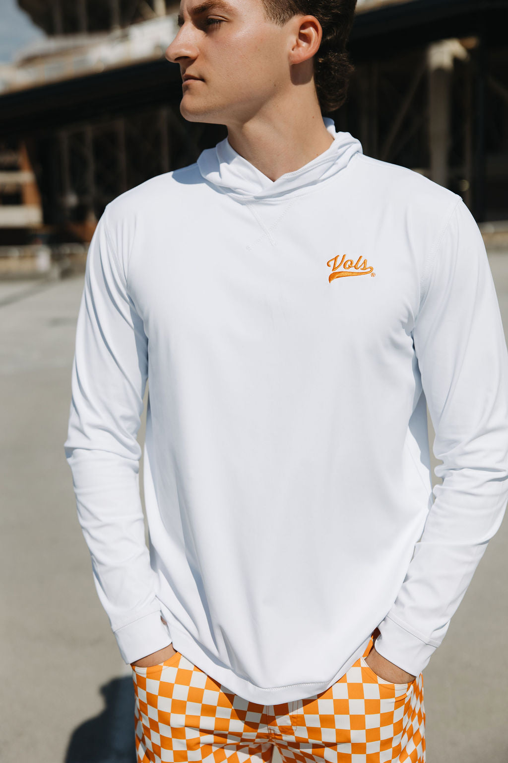 A person stands outdoors wearing a white long-sleeve hoodie with Vols written on the chest and orange-and-white checkered shorts, with hands in pockets. The background shows an industrial structure.