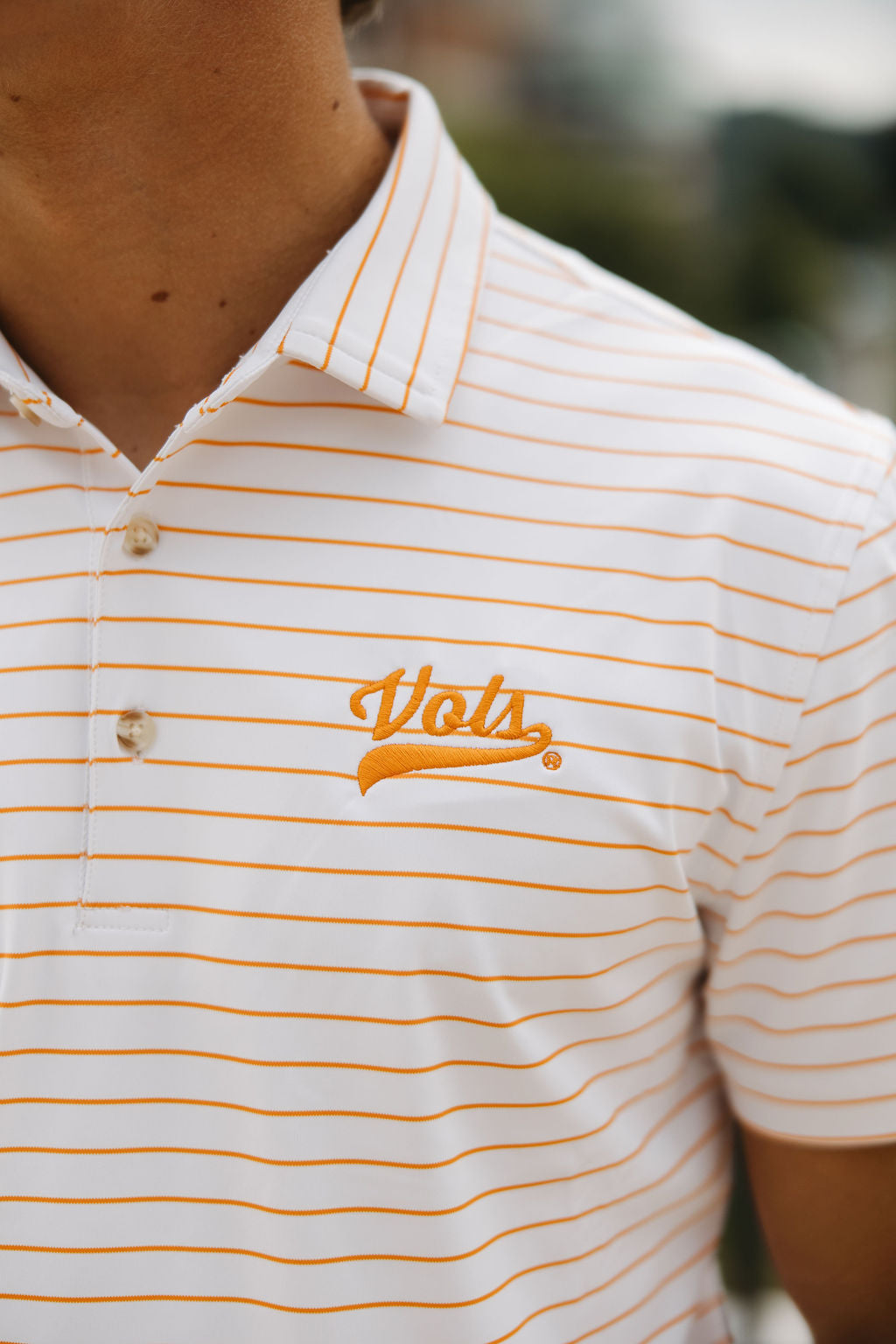 A person wearing a white polo shirt with thin orange horizontal stripes and an embroidered Vols logo in orange on the chest. The face is not visible.