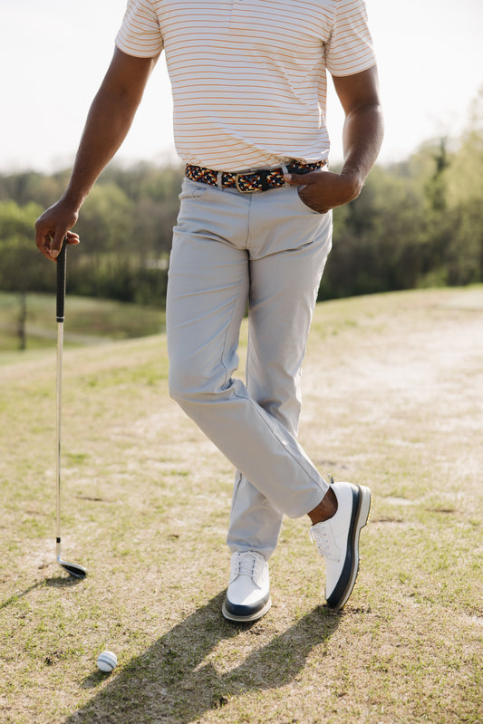 A man standing on a golf course with one hand in his pocket, holding a golf club and looking down at a golf ball on the ground. Only his body from neck down is visible. He wears light pants, white shoes, and a striped shirt.
