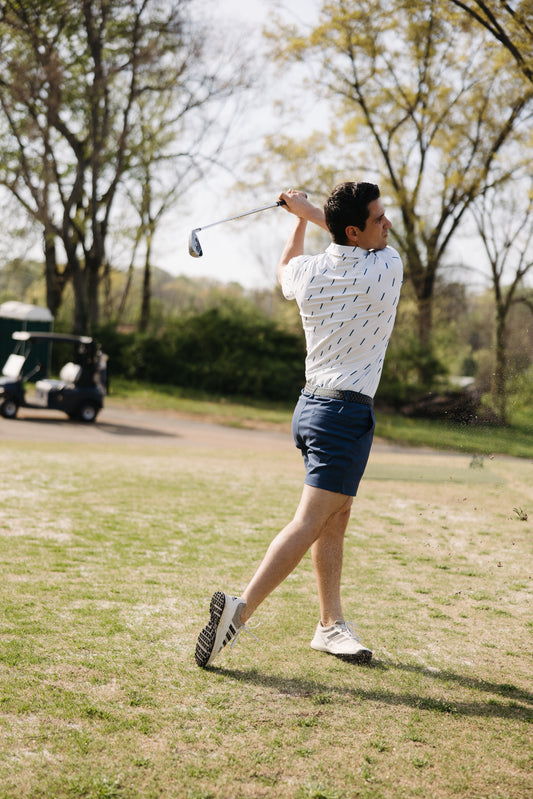 A man wearing a white patterned shirt and blue shorts follows through on a golf swing outdoors, with a golf cart and trees visible in the background on a sunny day.