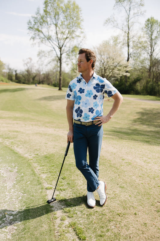 A man in a blue floral polo shirt and blue pants stands on a golf course holding a putter, with trees and grass in the background. He appears relaxed, looking off to the side.