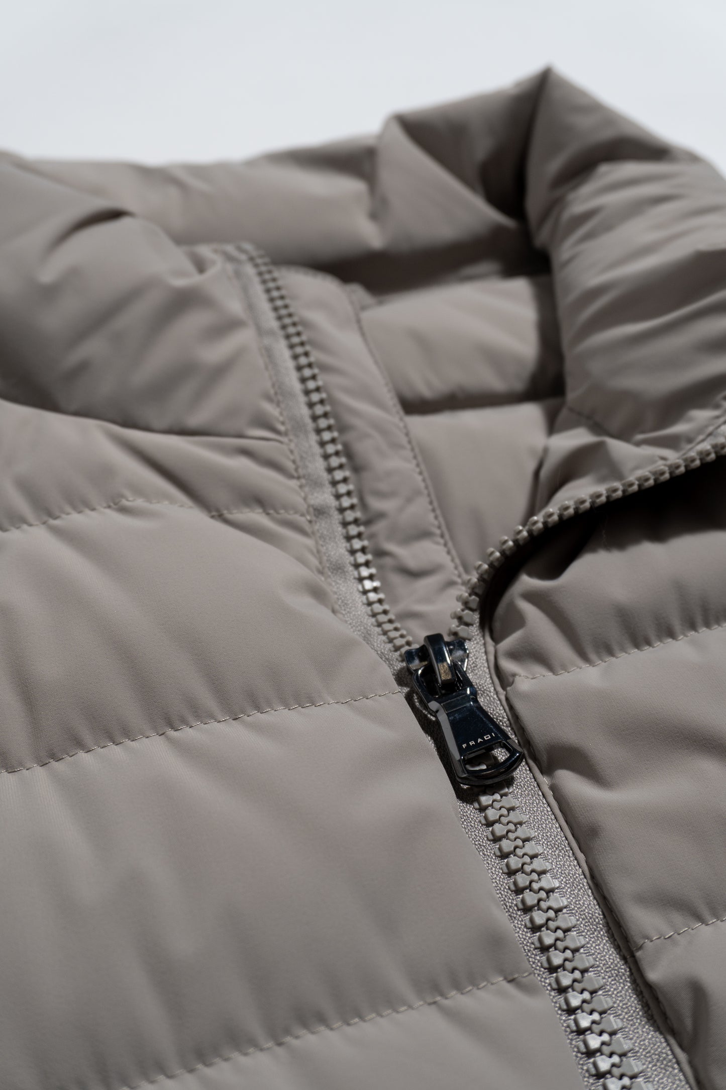 Close-up of a light gray, quilted puffer jacket with a prominent zipper partially unzipped. The fabric appears smooth and slightly shiny, emphasizing the jacket’s padded texture.
