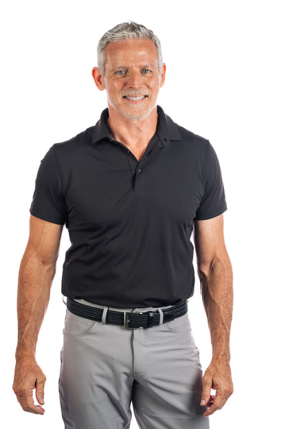 Photo of model smiling wearing a black golf polo and concrete five pocket pants on a white background.
