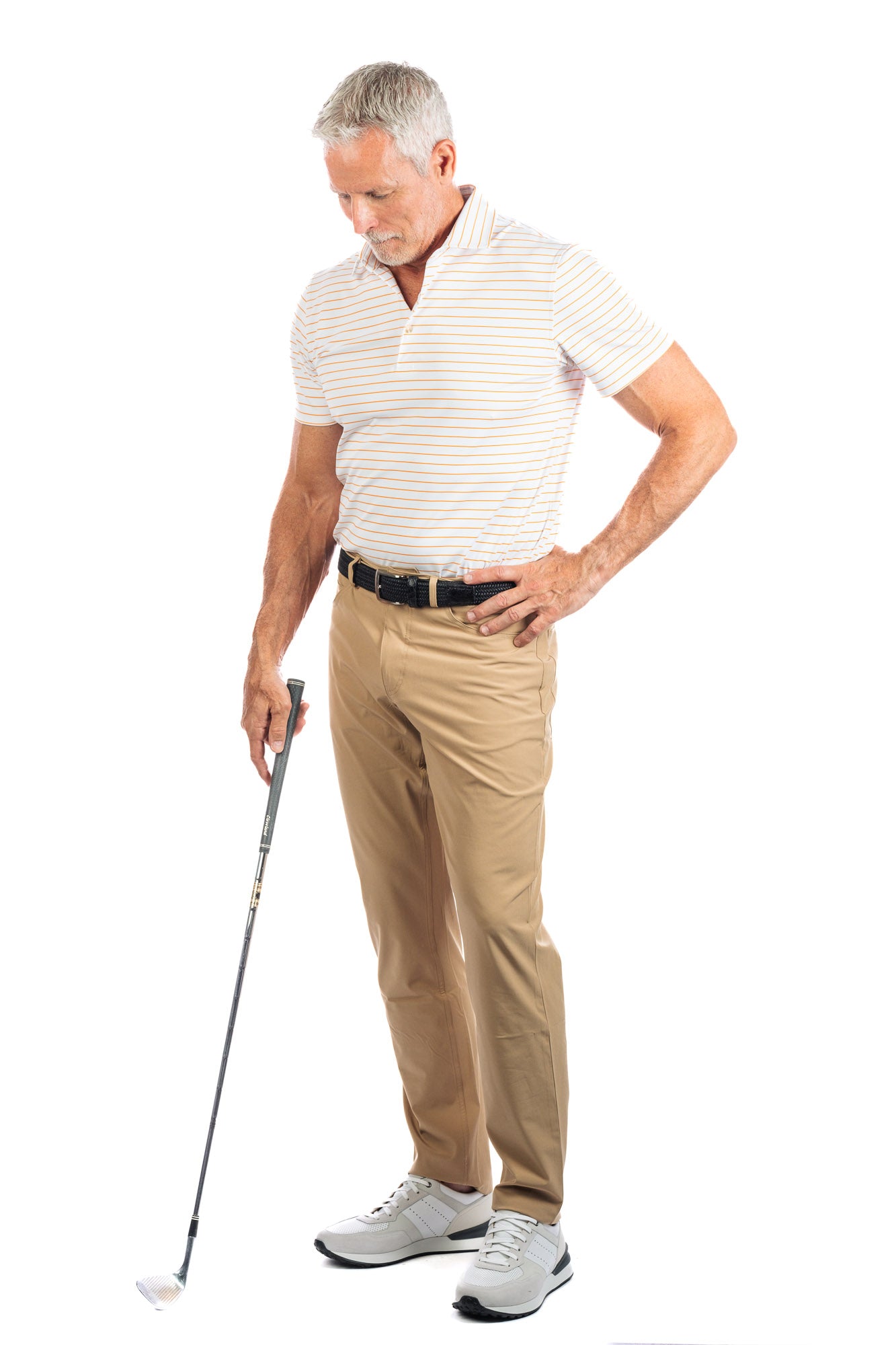 Full length photo of model wearing the Orange striped golf polo on a white background looking at the ground and holding a golf club. 