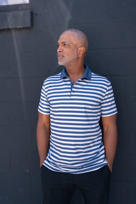 A man with short gray hair and a beard stands against a dark wall, wearing a blue and white striped polo shirt and black pants, with his hands in his pockets, looking thoughtfully to the side.